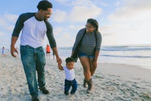 A dad and a mom happily walking along the beach in the sand with their 1 year old son.